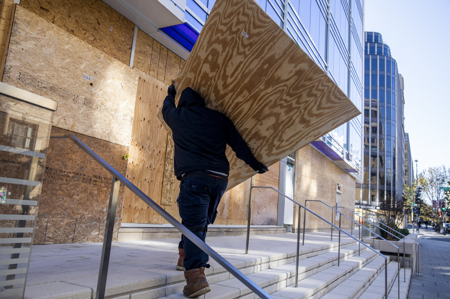 Một công nhân đang di chuyển một tấm ván gỗ lớn để gia cố cho cửa hàng tại một tòa nhà ở trung tâm thành phố Washington, để chuẩn bị cho các cuộc biểu tình có thể xảy ra. Cuộc biểu tình chống kỳ thị sau cái chết của George Floyd hồi tháng 5 khiến các cửa hàng thiệt hại nặng nề vì bị đập phá, cướp bóc - Ảnh: WAMU.