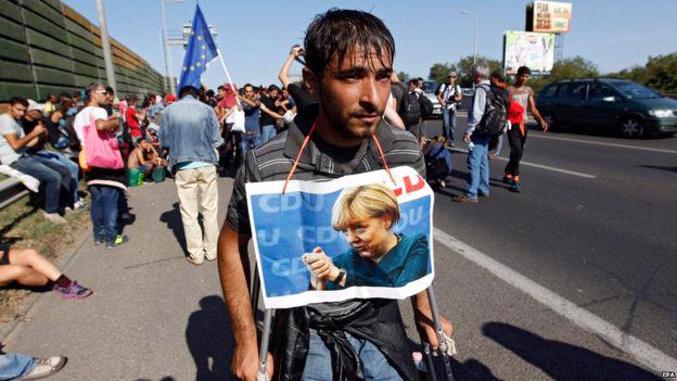 Tấm hình Thủ tướng Angela Merkel - người đã tạo điều kiện cho dòng người tị nạn đổ vào Đức năm qua - được đeo trên cổ một người nhập cư từ Trung Đông - Ảnh: BBC.<br>
