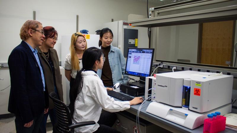 (Từ trái qua) Phó Giáo sư kỹ thuật hóa học Rong Tong; Phó Giáo sư khoa học và kỹ thuật vật liệu Wenjun "Rebecca" Cai; cùng hai sinh viên Eungyo Jang và Ziyu Huo họp bàn xung quanh Liqian Niu (ngồi), đang làm việc với máy Luminex 200 dùng để phân tích nồng độ cytokine của khối u. Ảnh: Hailey Wade từ Virginia Tech.